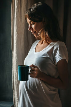 young pregnant woman looking out of a window drinking a cup of tea