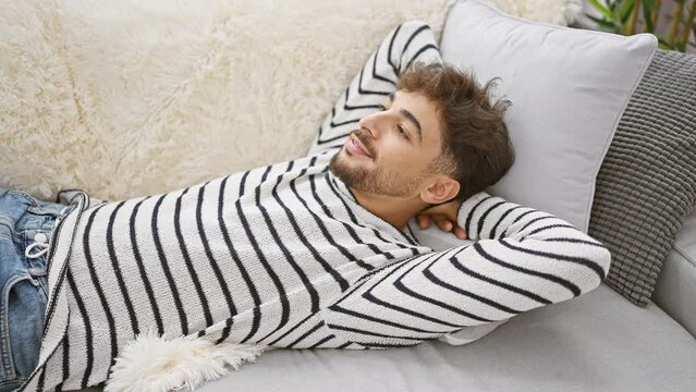 Heartwarming sight of a young, handsome arab man, full of joy and positive vibes, confidently lying on a cozy sofa at home, hands at ease behind his head, smiling, truly comfortable in his relaxation.