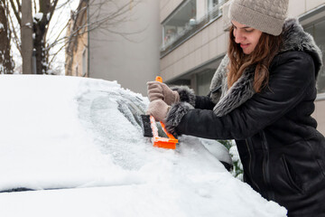 Young  woman using a snow brush to remove snow from her car's windshield. Concept of winter...