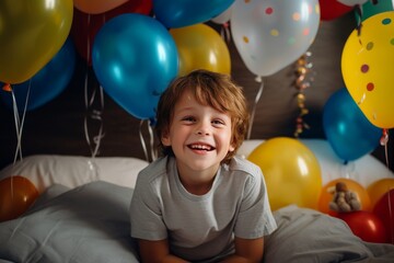 Fototapeta na wymiar A happy little boy is sitting on the bed in his room decorated with a lot of colorful balloons and smiling. The concept of holidays and happy birthday surprise. Generated by AI.