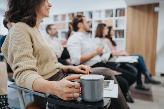 Selective focus on an engaged audience member holding a coffee mug at a seminar. Attendees in casual and formal wear attentively listen to a presentation, suggesting a professional development or