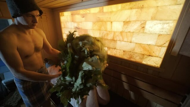 Man lying on wooden bench in SPA with masseur doing massage with oak broom. Client in Finn sauna indoors with professional.