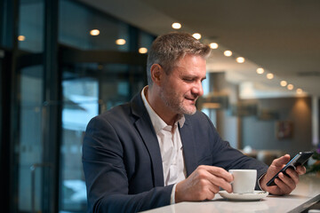 Adult focused caucasian businessman using smartphone while drinking tea