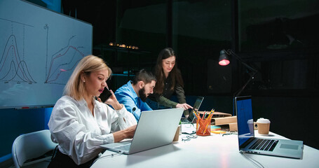 Caucasian young blonde woman talking on mobile phone and working at laptop computer in office at night shift. Male and female company co-workers cooperating on background. Lady speaking on cellphone.