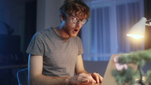 Portrait of amazed smiling young man got notification message and looking at screen with puzzled face at late night at home Shocked male reacting to news and feel confused next to laptop indoors