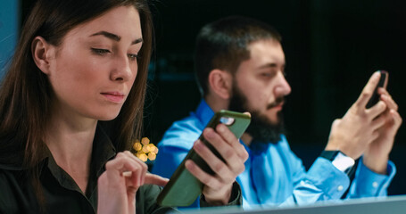 Caucasian man and woman sitting at office in evening at working place and tapping or texting on smartphones while female co-worker typing on laptop computer. Night shift work concept.
