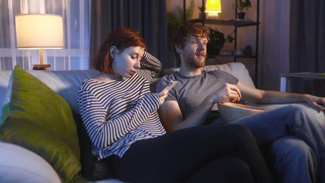 Boring young woman hold smartphone scrolling social media and ignoring her boyfriend or husband with popcorn laughing watching comedy movie show serial enjoying leisure time at home together at night