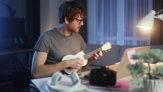 Handsome young red haired man watch video tutorial on ukulele guitar playing or having personal lesson with teacher distance remote on laptop computer late at home Online musical education concept 