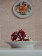 Vertical table top close up view of an open pomegranate on a beige plate against the white table and pastel pink wall with a rosemailing painted cutting board on it.