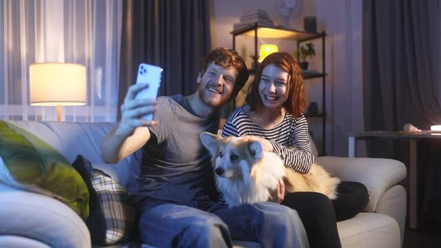 Portrait of young red hair couple with corgi dog pet grimacing taking selfie photograph saving great memories on smartphone mobile phone at home at night Happy relationship concept