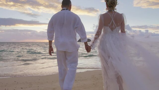 Attractive couple in boho style clothes raising up hands celebrating the best moment of life. Happy excited couple in love getting married, holding hands and walking barefoot by sandy Hawaiian beach