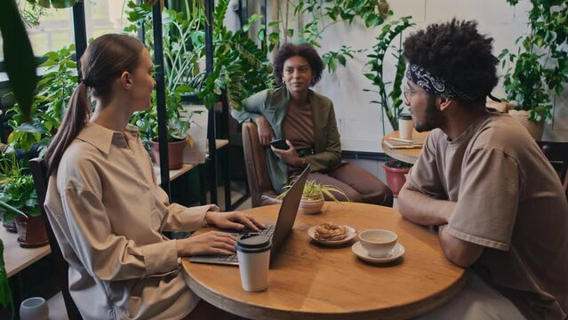 Medium shot of Caucasian woman with laptop and biracial man sitting at table in office canteen or coworking and chatting to female African American colleague at next table