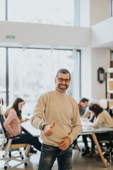 Fototapeta na wymiar Cheerful male in casual attire giving a thumbs up sign with a beaming smile in a vibrant office setting, team working in the background.