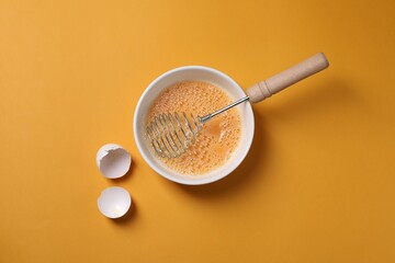 Beaten eggs, metal whisk in bowl and shells on orange background, flat lay