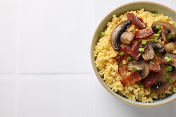 Tasty millet porridge with mushrooms, bacon and green onion in bowl on white tiled table, top view. Space for text