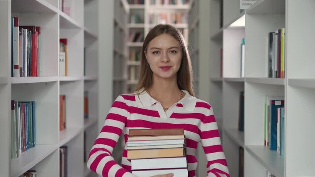 Happy female student holds books in aisle between bookcases. Blonde woman carries literature for reading in modern bookstore. Lady with folio collection