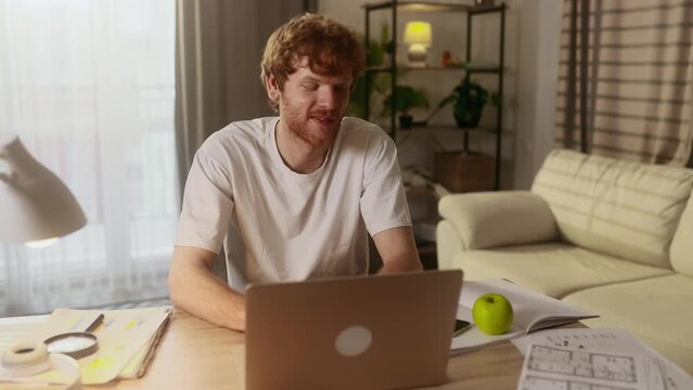 Handsome young red haired man working on laptop computer having distance remote work or study and eating fresh red apple at home workplace Healthy food concept