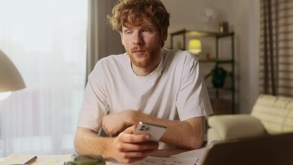 Boring young red haired man scrolling feed of social networks products in internet store on smartphone while take a break indoors Tired exhausted male look at screen without enthusiasm at home