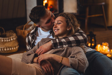 Beautiful young loving couple relaxing near fireplace in bed at home. Happy spouses enjoying lazy romantic winter cozy morning in bedroom. Candles, garlands, countryside interior. Romantic date - obrazy, fototapety, plakaty
