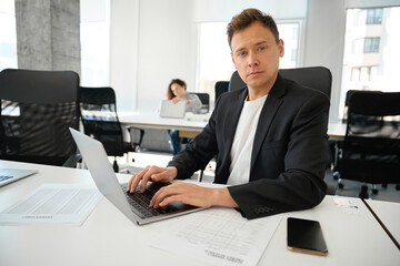 Self-confident man looking at camera and working on laptop