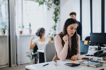 Fototapeta premium Focused female professional working at her desk with colleagues in background in a modern office setting.