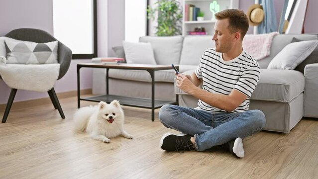 Handsome young caucasian man sitting on living room floor, smiling confidently, makes a snap of his adorable dog with smartphone at home