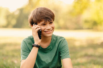 Smiling handsome teen boy has a phone talk with his friend while sitting in park.