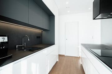 This is a black and white kitchen with a sink, stove, and door. The countertops are granite-like, and the appliances are also black colored. The walls are painted white, floor is made of wood. 