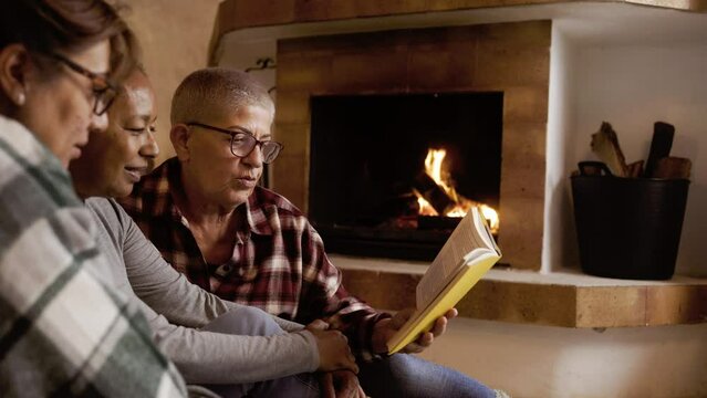 Multiracial senior friends reading book by fireplace inside rural home