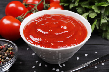 Delicious ketchup in bowl and spices on black wooden table, closeup. Tomato sauce