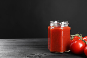 Delicious ketchup in jar and tomatoes on black wooden table, closeup with space for text. Tomato sauce