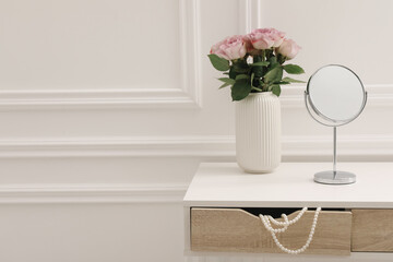 Mirror and vase with pink roses on white dressing table in makeup room, space for text