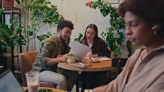 Medium selective focus shot of Caucasian woman and Arab man sitting together at table in cozy cafe, drinking coffee and looking through business documents, and black woman working on laptopMedium sele