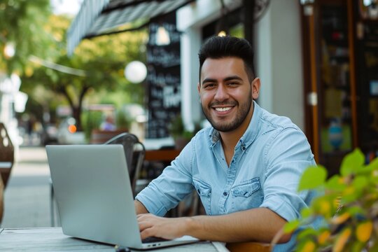 Happy Young Latin Business Man Using Laptop Sitting Outdoors. Smiling Hispanic Guy Student Or Professional Looking At Computer Sitting In City Cafe Elearning Or Hybrid Working, Searching Job Online