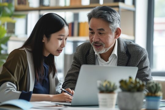 Mid aged Latin male manager mentor teaching young Asian female worker looking at laptop discussing corporate strategy in teamwork, working on computer in office at international team meeting