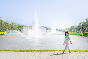 Asian Girl Strolling by Park Fountain on Sunny Day, Kid in dotted outfit takes leisurely stroll by...