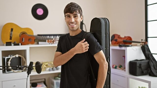 Charming young hispanic man ecstatic with confidence, showcasing his guitar case in music studio, all set to rock the concert