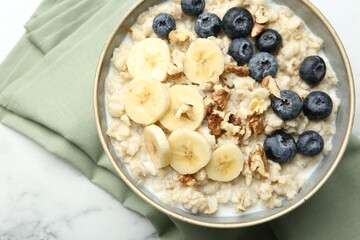 Tasty oatmeal with banana, blueberries, walnuts and milk served in bowl on white marble table, top view