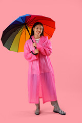 Beautiful young Asian woman in raincoat with umbrella on pink background