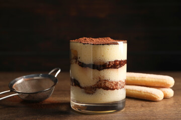 Delicious tiramisu in glass, biscuits and sieve on wooden table, closeup