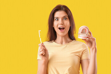 Surprised young woman with dental braces holding toothbrush and jaw model on yellow background
