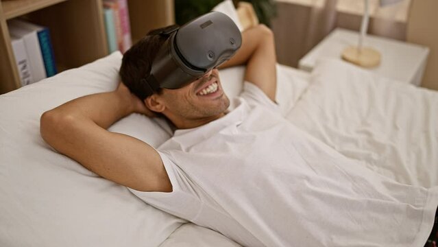 Happy youthful hispanic guy captivated by online game, enjoying futuristic vr headset, lying in bedroom with gadget, confident in his gaming ability, indoor enthusiast with radiant smile.