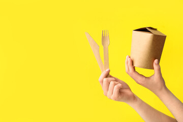 Female hands with takeaway paper box and cutlery on yellow background - obrazy, fototapety, plakaty