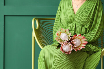 Young woman with beautiful protea flowers sitting in armchair on green background, closeup