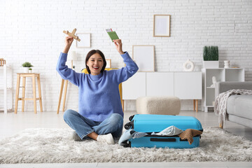 Young woman with wooden plane, passport and suitcase sitting on floor at home