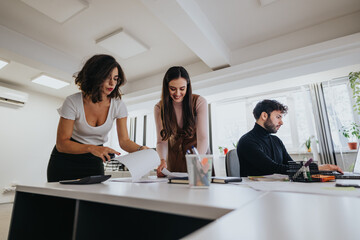 Team collaboration in modern office with two women discussing over papers and man working on laptop.