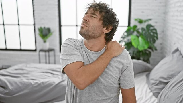 An attractive man with a beard grimaces in pain, clutching his shoulder in a modern bedroom setting.