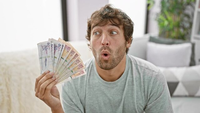 Shocked young man at home, mouth wide open in disbelief, holding colombian pesos. amazingly scared expression painted on his face!