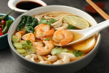 Eating delicious ramen with shrimps from bowl with spoon at grey table, closeup. Noodle soup