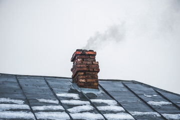 Smoke comes from the chimney of the house with a tin roof in winter. Chimney in red bricks on the...